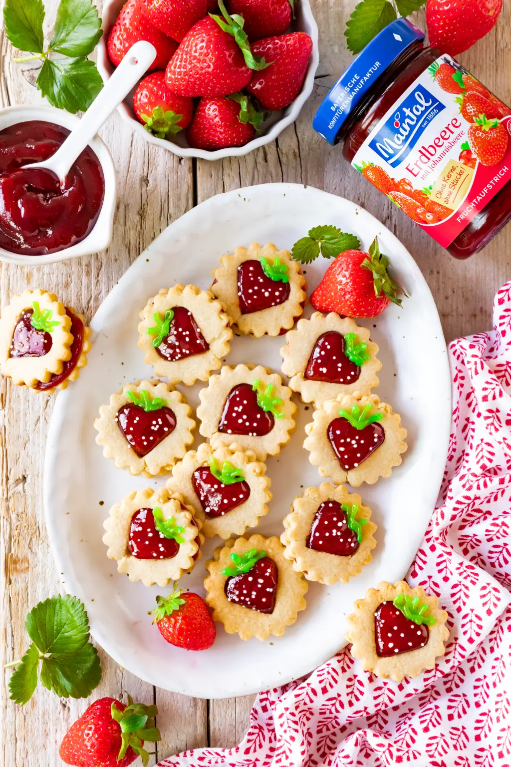 Kekse in Erdbeeroptik mit roter Füllung auf weißer Platte, dekoriert mit grüner Zuckerschrift und Zuckerperlen, umgeben von Erdbeeren und Fruchtaufstrich.