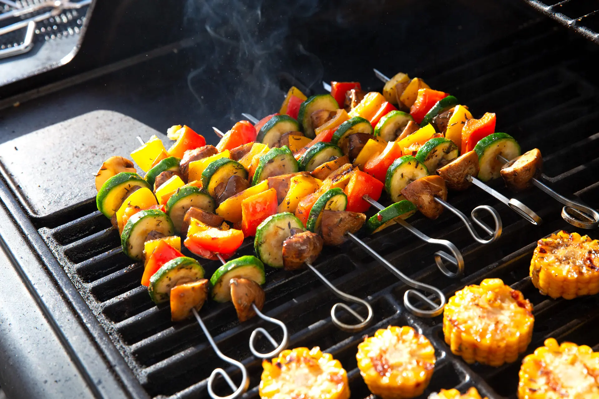 Bunte Gemüsespieße mit Paprika, Zucchini und Champignons grillen auf Rost, daneben Maiskolbenstücke