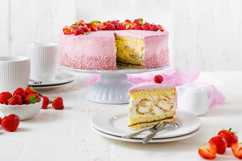 Serviervorschlag Windbeuteltorte mit Gartenfrüchten auf Tortenplatte, Teller mit einem Stück der Torte, Kaffeetassen und Schüssel mit frischen Erdbeeren.