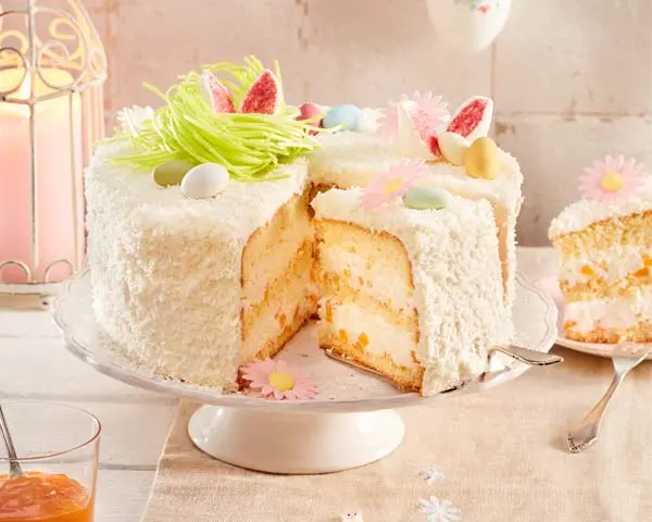 Aprikosen-Kokos-Ostertorte mit Deko Aprikosen-Kokos-Ostertorte auf Tortenplatte serviert, daneben ein Stück auf einem Teller.