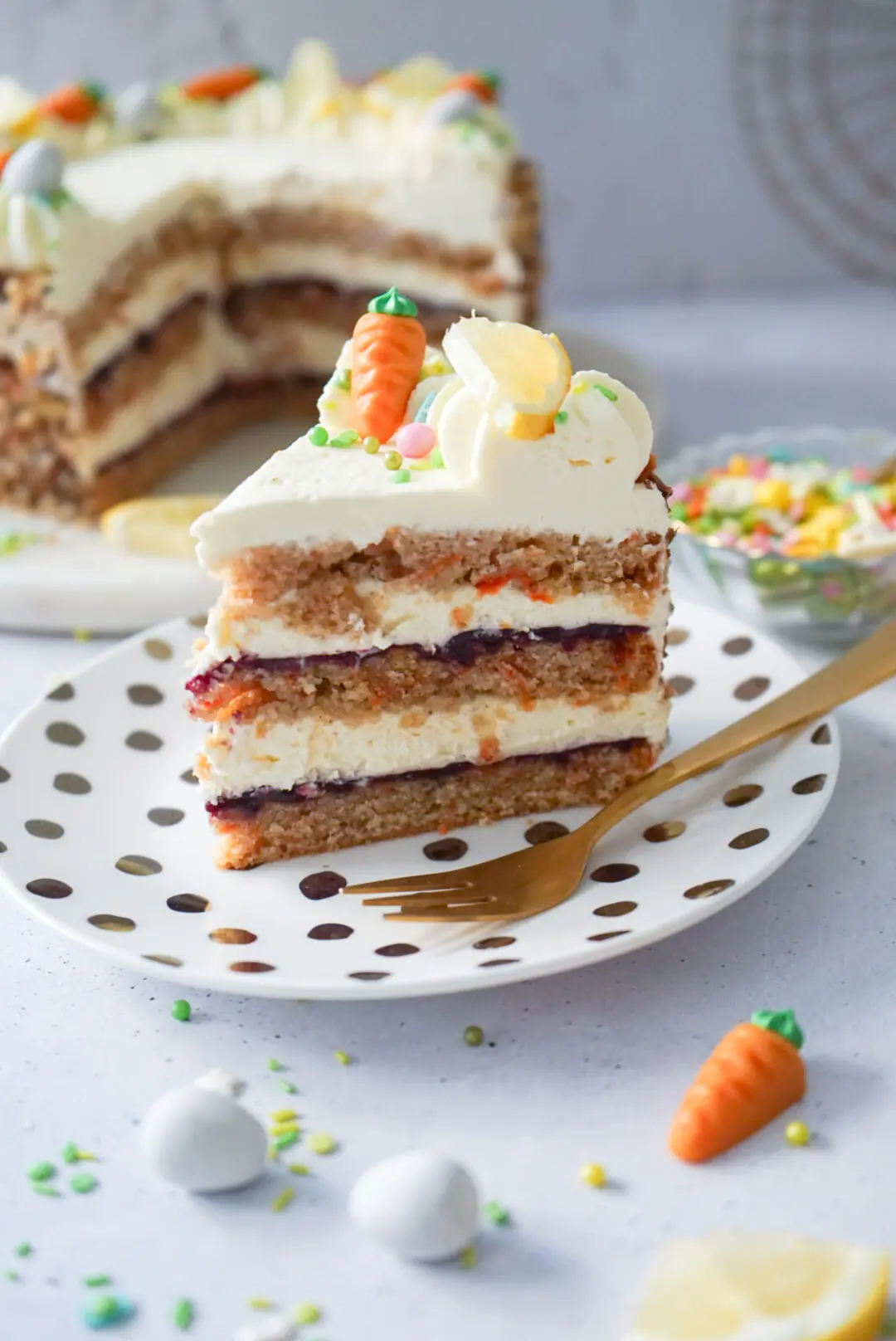 Rüblitorte mit Zitronencreme und Waldbeeren Fruchtaufstrich, dekoriert mit Marzipan-Karotte auf einem Teller.