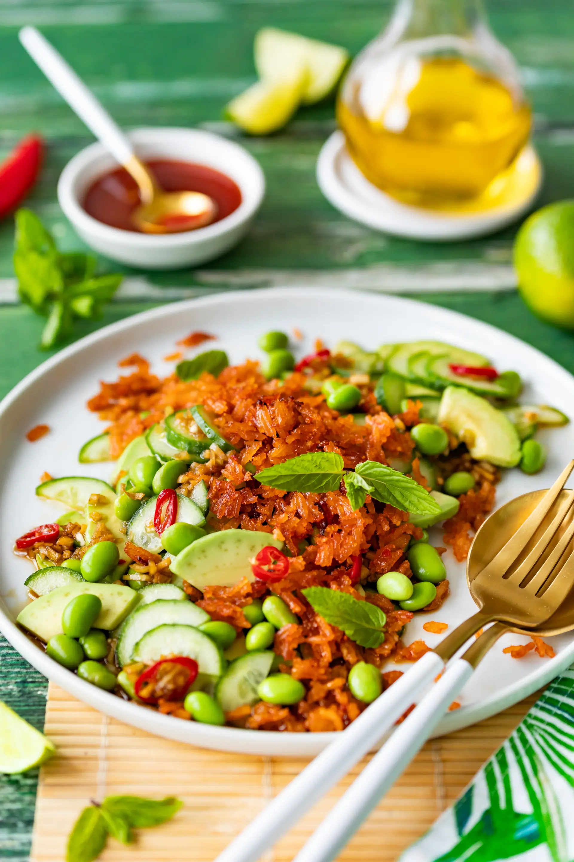 Crispy Reis Salat mit Gurke, Avocado, Edamame und Minze auf einem weißen Teller, garniert mit Chilischoten. Im Hintergrund Ketchup und Olivenöl.