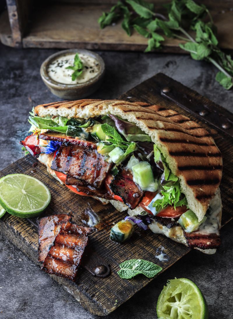 Serviervorschlag Veganer Grill-Döner mit Tofu-Bacon: auf Holzbrett mit Limettenscheiben, KOriander, Schälchen mit süß-sauer Dressing.