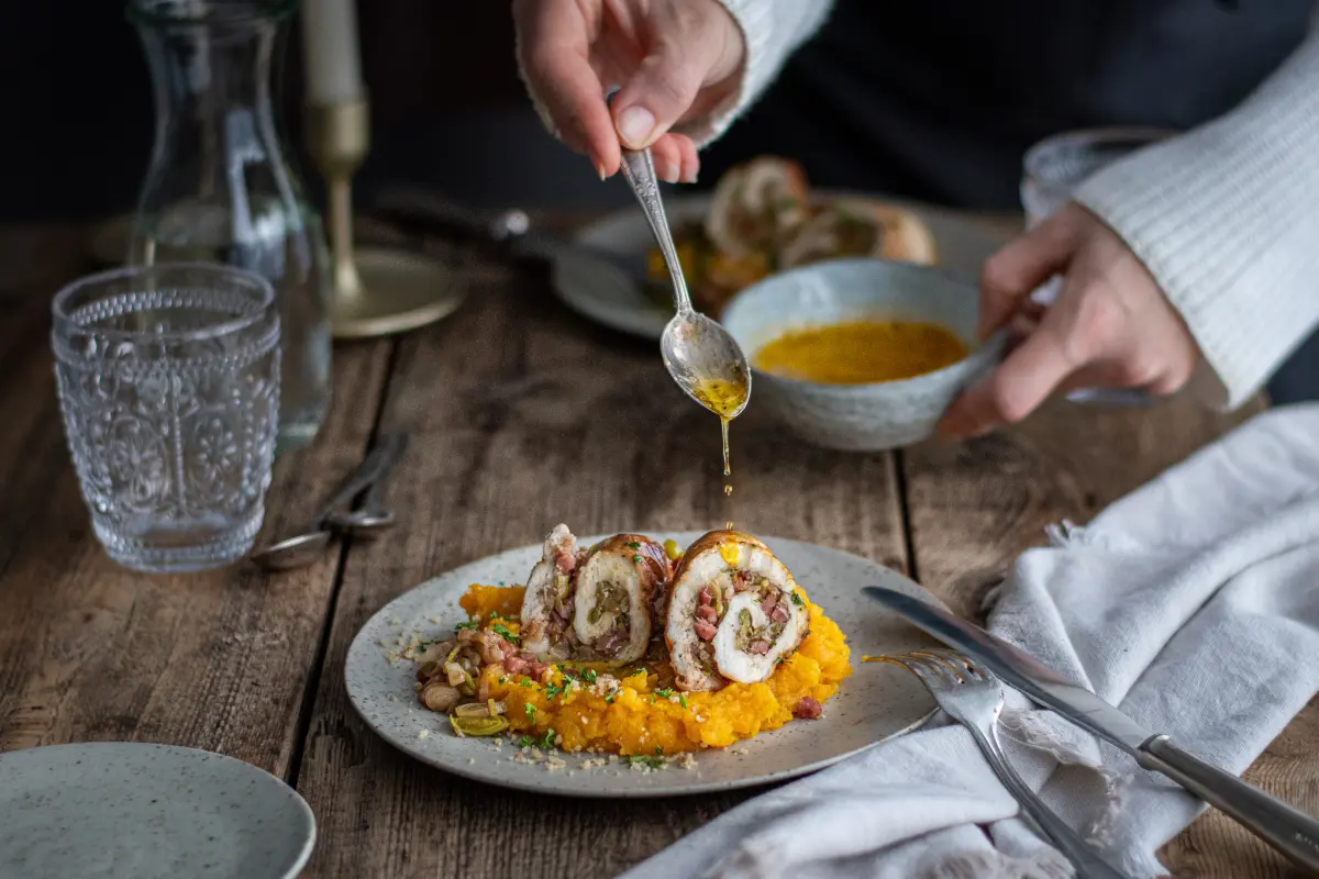 Gedeckter Tisch mit Teller von gefülltem Hähnchenröllchen mit Kürbis-Kartoffelbrei, Glas, Karaffe. Über das Gericht wird Knoblauchbutter geträufelt.