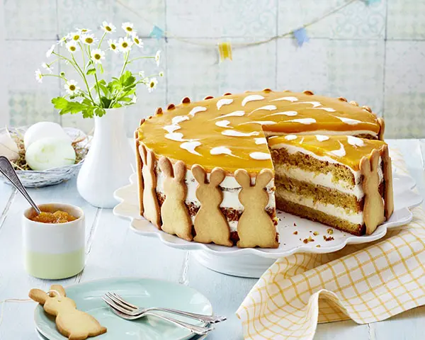 Kaffeetafel mit Karotten-Aprikosentorte mit Osterhasenkeksen auf Tortenplatte, Teller mit Osterhasenkeks, Schüssel mit Aufstrich, Blumen uns Osternest.
