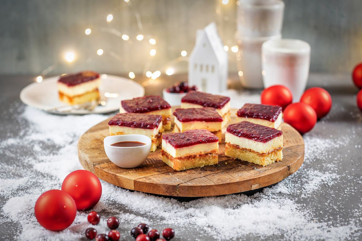 In Stuecke geschnittener Blechkuchen mit Preiselbeer-Gluehweinspiegel auf einer Holzplatte, Teller mit Kuchenstück, Kugeln und winterlicher Deko.