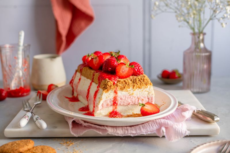 No Bake - Sahne-Quarkcreme-Torte mit Erdbeeren Serviervorschlag No Bake - Sahne-Quarkcreme-Torte mit Erdbeeren in tieferem Teller auf Platte mit Glas Erdbeersauce, Vase mit Blumen.