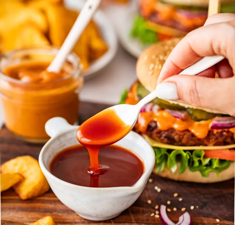 Veggie Cheeseburger mit Hagebutten-Soße Löffel mit fruchtigem Hagebutten-Chutney über einer Schale, daneben ein vegetarischer Cheeseburger auf einem Holzbrett mit Kartoffelspalten und einem Glas Dip im Hintergrund.